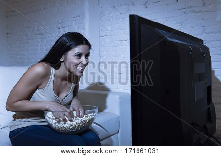 young beautiful latin woman home alone watching television smiling sitting at sofa couch on living room happy and excited enjoying TV show or movie relaxed in dark light eating popcorn