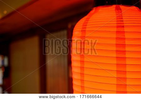 Close-up detail of the top half of a capsule shaped paper lantern shining with an orange light with traditional Japanese architecture in the background. Travel and culture concept.