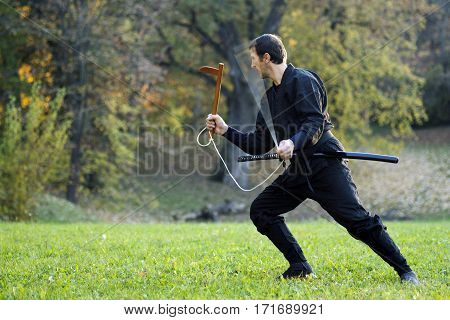 Ninja in black kimono is practicing with kusarigama outdoors in the park.