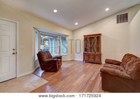 Interior of a rambler home features vaulted ceiling over yellow walls framing a bay window. Entrance room furnished with brown sofa and vintage cupboard atop bamboo wood floors. Northwest USA