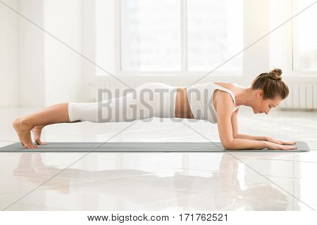 Young attractive yogi woman practicing yoga, standing in Dolphin Plank exercise, phalankasana pose, working out, wearing sportswear, sport bra, pants, indoor full length, white color room background