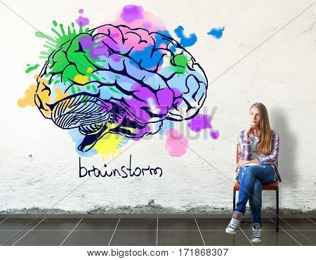 Thoughful european girl sitting on chair in concrete room with bright human brain sketch on wall. Creative thinking concept. 3D Rendering