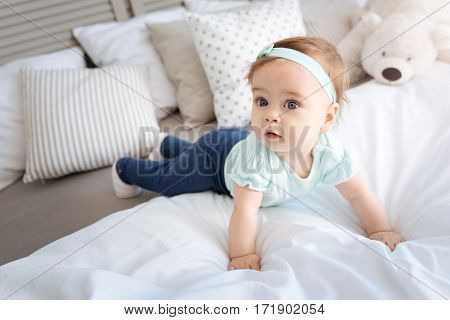Exploring this world. Cute curious little girl lying on the bed in the bedroom and looking away while expressing interest