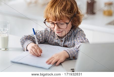 Little genius. Persistent lively ginger kid carefully writing down his thoughts while wearing big glasses and sitting at white table