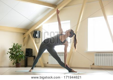 Young attractive woman practicing yoga, standing in extended triangle exercise, Utthita Trikonasana pose, working out, wearing sportswear, grey tank top, pants, indoor full length, home interior