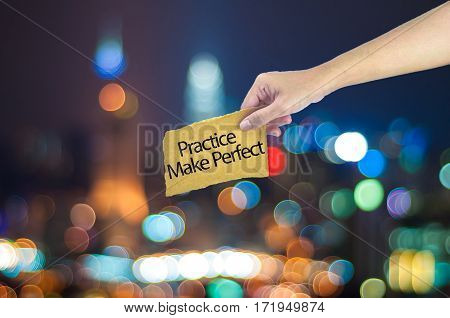 Hand Holding A Practice Make Perfect Sign Made On Sugar Paper With City Light Bokeh As Background