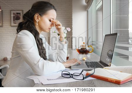 Elegant lady holding nose with closed eyes because of stress and migraine at work. Person tired of work sitting at table with laptop, papers, tea, glasses and folders. Exhausted woman at workplace