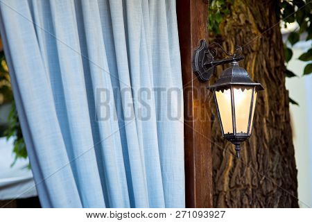 A Street Lamp On A Brown Pillar Against The Background Of Blue Curtains And A Tree Trunk, An Antique