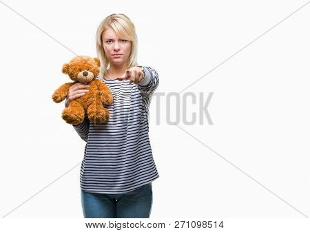 Young beautiful blonde woman holding teddy bear plush over isolated background pointing with finger to the camera and to you, hand sign, positive and confident gesture from the front