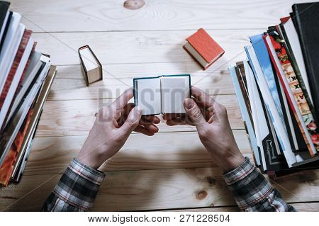 A Man Reading A Miniature Book Among The Big Books In The Library At The Wooden Table.