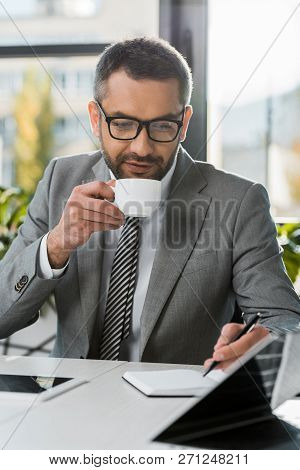 Businessman In Suit And Eyeglasses Drinking Coffee And Writing In Notebook At Wokrplace