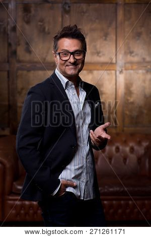 Emotional Happy Man Standing With Brown Sofa Behind His Back And Using Gestures While Posing For A P