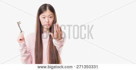 Young Chinese real state agent woman over isolated background holding vintage key with open hand doing stop sign with serious and confident expression, defense gesture
