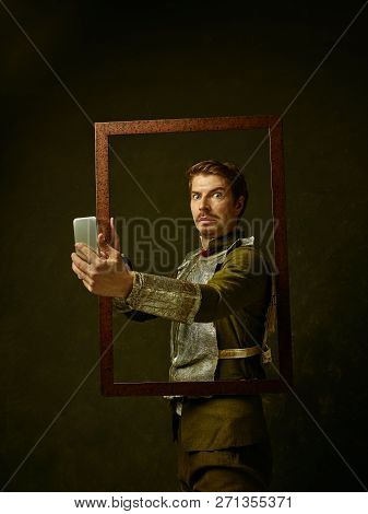Medieval Knight With Mobile Phone On Dark Studio Background. Portrait In Low Key Of Brutal Man In Tr
