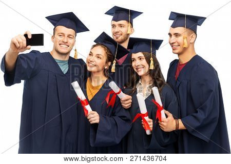 education, graduation and people concept - group of happy graduate students in mortar boards and bachelor gowns with diplomas taking selfie by smartphone over white background