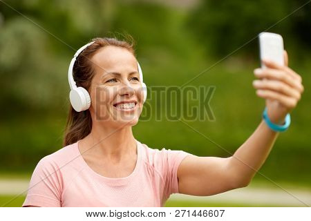 fitness, technology and healthy lifestyle concept - smiling woman in headphones listening to music and taking selfie by smartphone at summer park