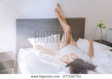 Young Woman Is Lying In Bed With Legs Up On Wall, Sunny Bedroom, White Bed Crumpled. Concept Weekend