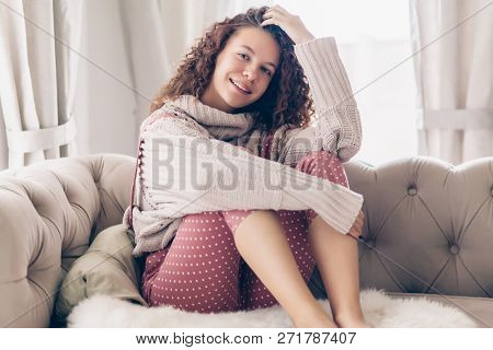 Hipster teenage girl with curly hair wearing beige knitted sweater and pink polka dot jumpsuit relaxing on a couch indoor