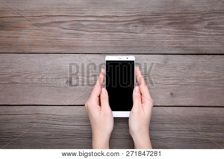Female Hands Holding Mobile Phone With Blank Screen On Grey Wooden Table. Smartfone White