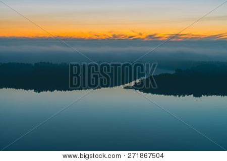 Early Blue Sky Reflected In River Water. Riverbank With Forest Under Predawn Sky. Yellow Stripe In P