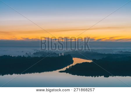 Early Blue Sky Reflected In River Water. Riverbank With Forest Under Predawn Sky. Yellow Stripe In P