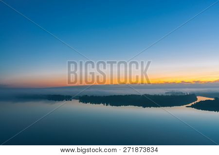 Broad River Flows Along Diagonal Shore With Forest Under Thick Fog. Early Blue Sky Reflected In Wate