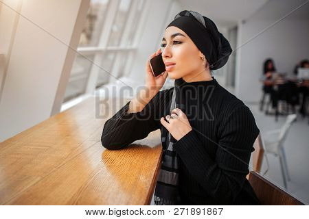 Cheerful Young Arabian Woman Sit At Tall Table And Look At Window. She Talks On Phone. Moel Touches 