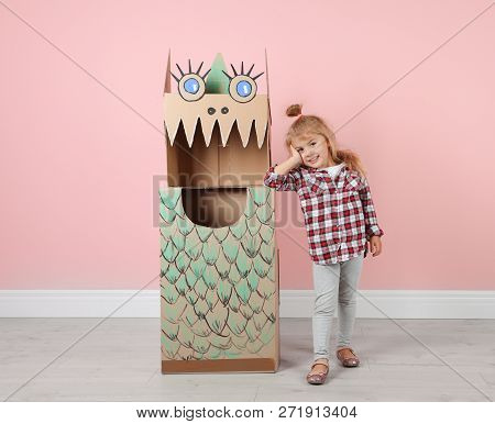 Little Girl And Cardboard Costume Of Dinosaur Near Color Wall Indoors. Space For Text