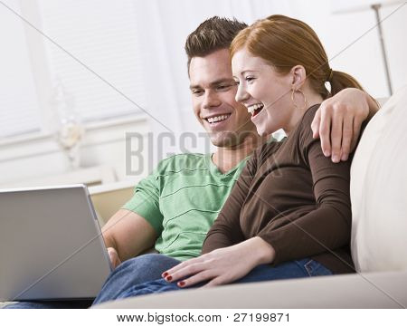 A young and attractive couple sitting together and viewing a laptop screen. They are smiling and laughing. Horizontally framed photo.