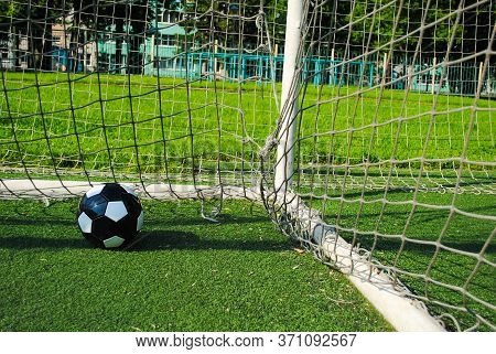 Black Soccer Ball In Goal Net On A Green Lawn On A Sunny Summer Day. The Goal Net Is Dirty And Torn.