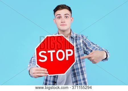 Young Man Is Holding Red Prohibition Road Sign Stop On Blue Background. Serious Guy Is Warning About