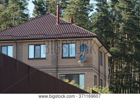 Part Of A Large Private Brown Brick House With Windows And Black Satellite Dishes Under A Tiled Roof