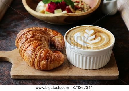 A cup of coffee with latte art on top, butter  croissant and granola with fresh tropical fruits and 