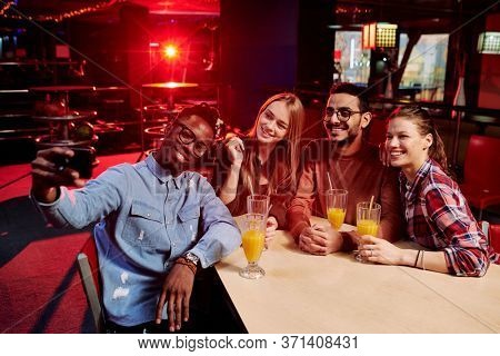 Four happy friends in casualwear making selfie by table in the bar while having drinks after watching bowling game of their favorite team