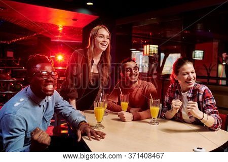 Young cheerful fans of various ethnicities watching broadcast of their favorite bowling team while sitting by table and having drinks