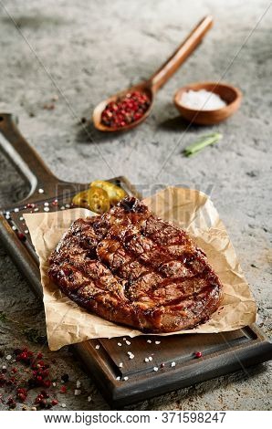 Classic ribeye steak served in waxed paper on wooden cutting board. Homemade barbecue dish concept. Roasted beef steak with yellow chilli pepper slices and salt crumbs. Rustic spoon and spice bowl