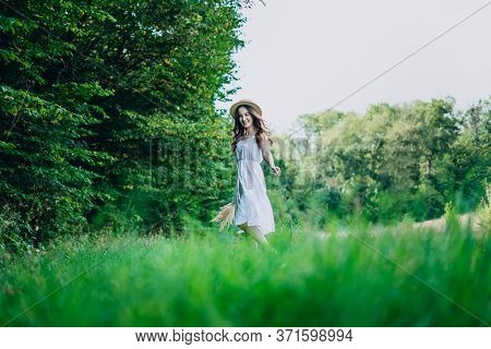Young Woman Walking In A Green Field. Young Girl In A White Dress And Hat. Girl In A Dress And A Sum