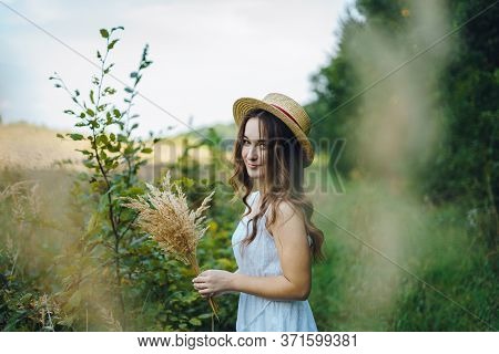 Smiling Girl In A Straw Hat Collects Flowers. Girl In A Summer Dress In The Tall Grass. Woman In A G