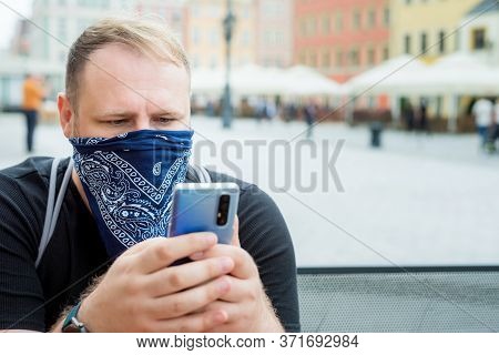 Adult Man In Bandana As A Face Mask Uses Smartphone In City Cafe Terrace. Solo Outdoor Activities. S