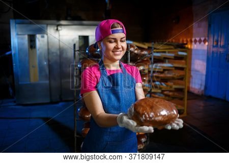 A Baker Girl Takes Hot Bread In A Bakery Against The Background Of Shelving With Bread. Industrial P