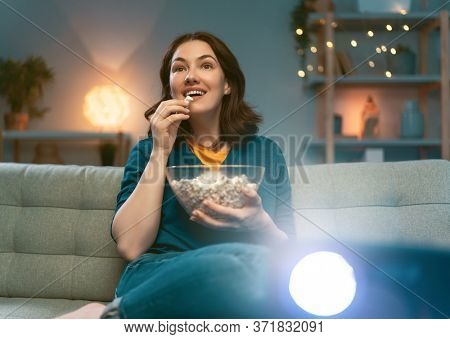 Young woman watching projector, TV, movies with popcorn in the evening. Girl spending time at home.