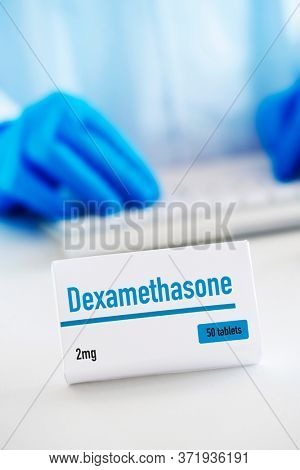 closeup of a simulated box of dexamethasone on a white table, and a man of a man wearing a white coat and blue surgical gloves in the background