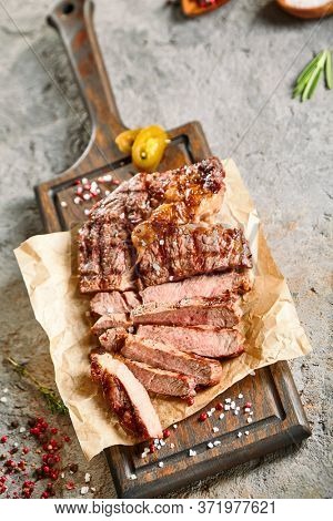 Classic ribeye steak served in waxed paper on wooden cutting board. Cut against grain beef steak. Roasted beef steak with yellow chilli pepper slices and salt crumbs. Rustic spoon and spice bowl
