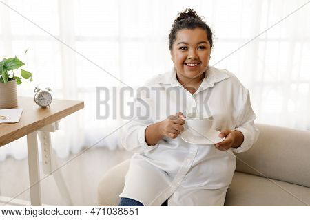 Positive Young Chubby Woman Sitting At Workdesk, Drinking Coffee
