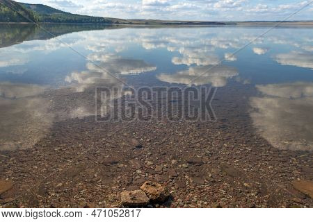 Lake Surrounded By A Rocky Shore, Mountains With Forest On Background.reflection Of White Clouds In 