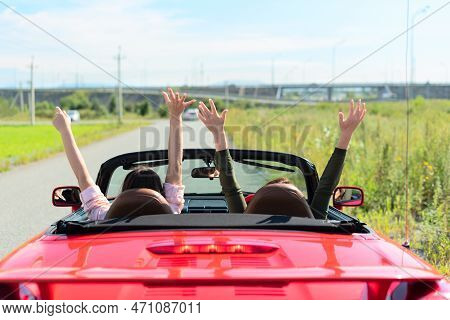 Back View Of Happy Girls Driving Red Cabriolet Car With Raised Hands During Vacation Road Trip Havin