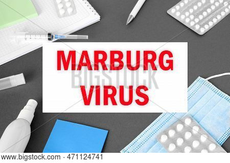 The Word Marburgvirus On Gray Modern Doctor Desk Table Background. Mask, Notepad, Syringe, Sanitizer