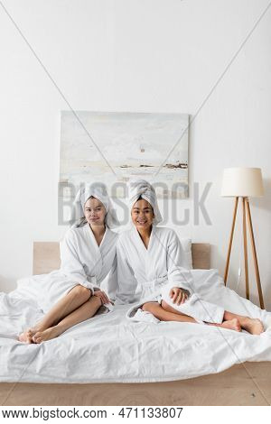 Full Length Of Young Barefoot Women In White Soft Robes And Towels Sitting On Bed And Smiling At Cam
