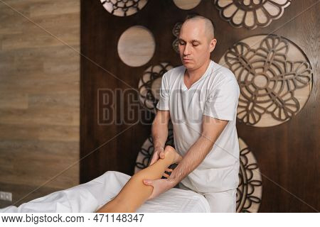 Serious Bald Male Masseur Massaging Lower Part Of Leg To Young Unrecognizable Woman In Spa Salon. Pr