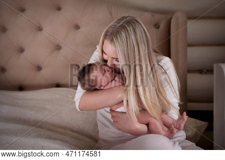 Mother Kisses Newborn Baby In Her Arms, Sitting On Bed. Beautiful Blonde Mom With Long Hair And Her 